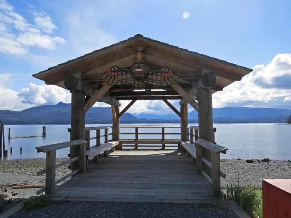 Wooden shelter on Alert Bay beach, with intricately carved eagle artwork above entrance. The calm ocean and forested mountains form the background