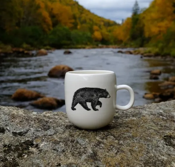 White mug with hand drawn bear image on front, set on rock in front of river surrounded by autumn foliage