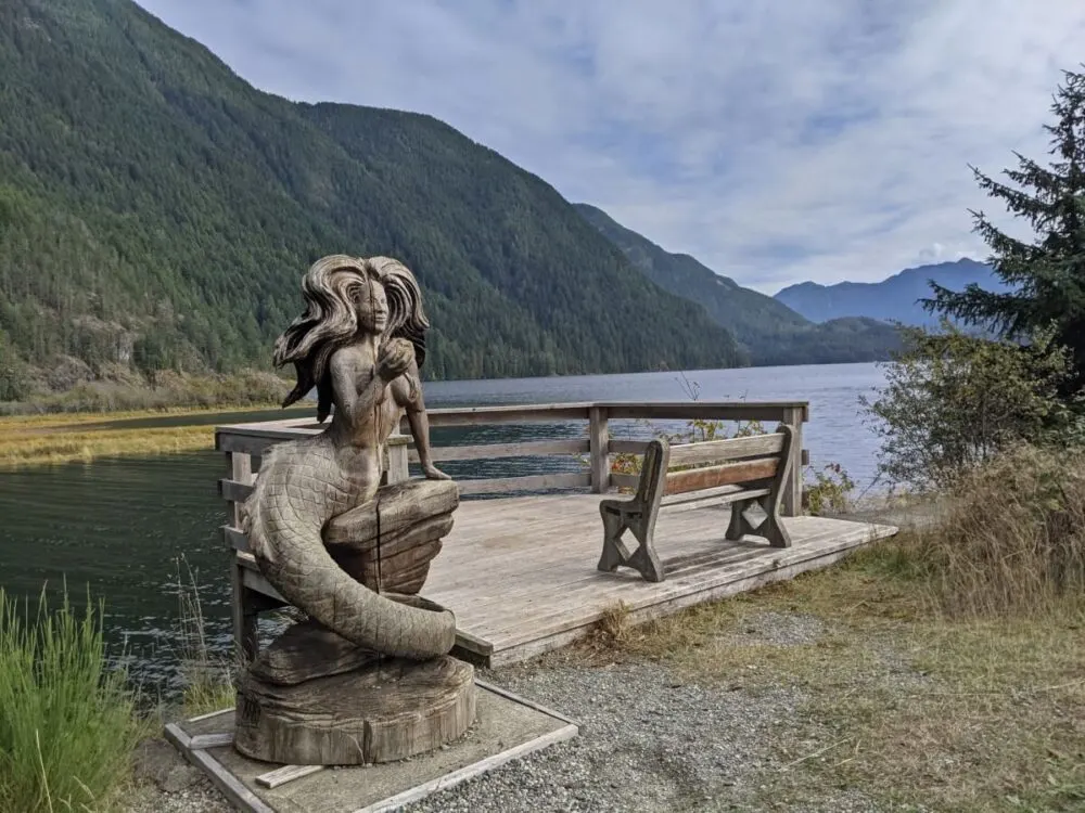 Carved wooden mermaid statue next to wooden bench on deck next to Gold River mouth, backdropped by forested mountains