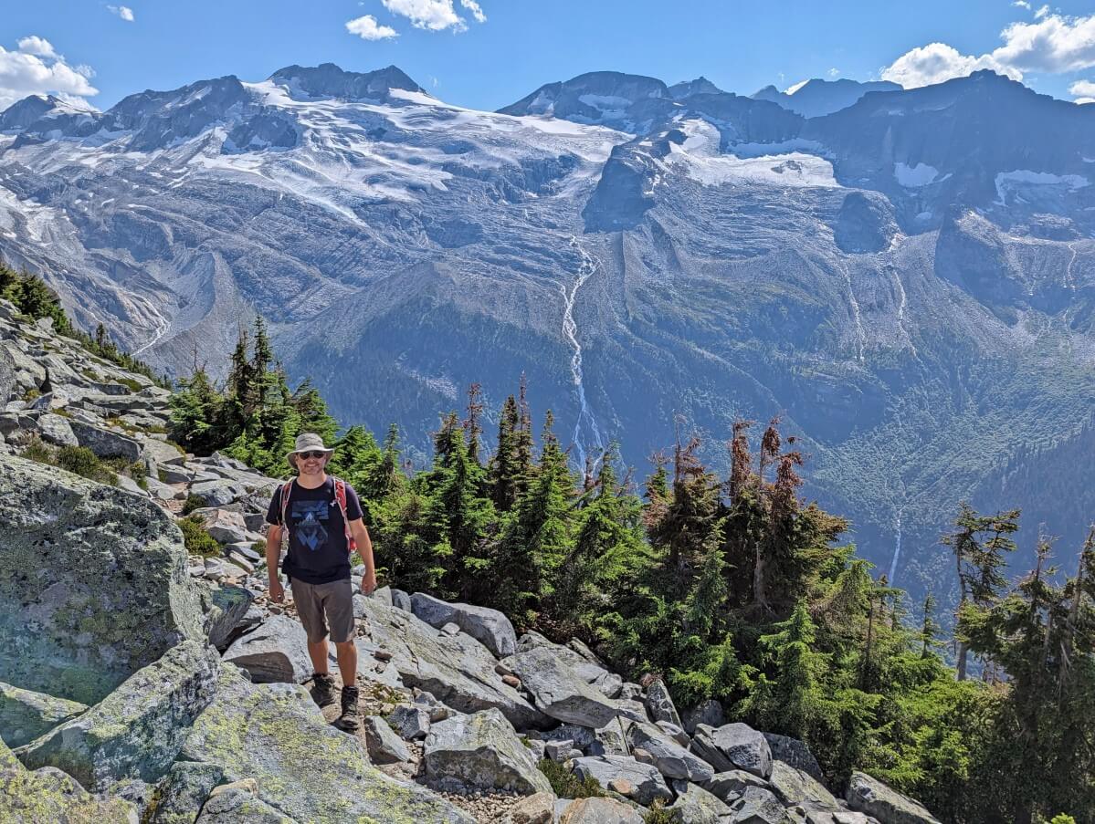 JR standing to camera in front of Asulkan Glacier, a large glacier at the peak of a mountain with waterfalls cascading down into forested valley below