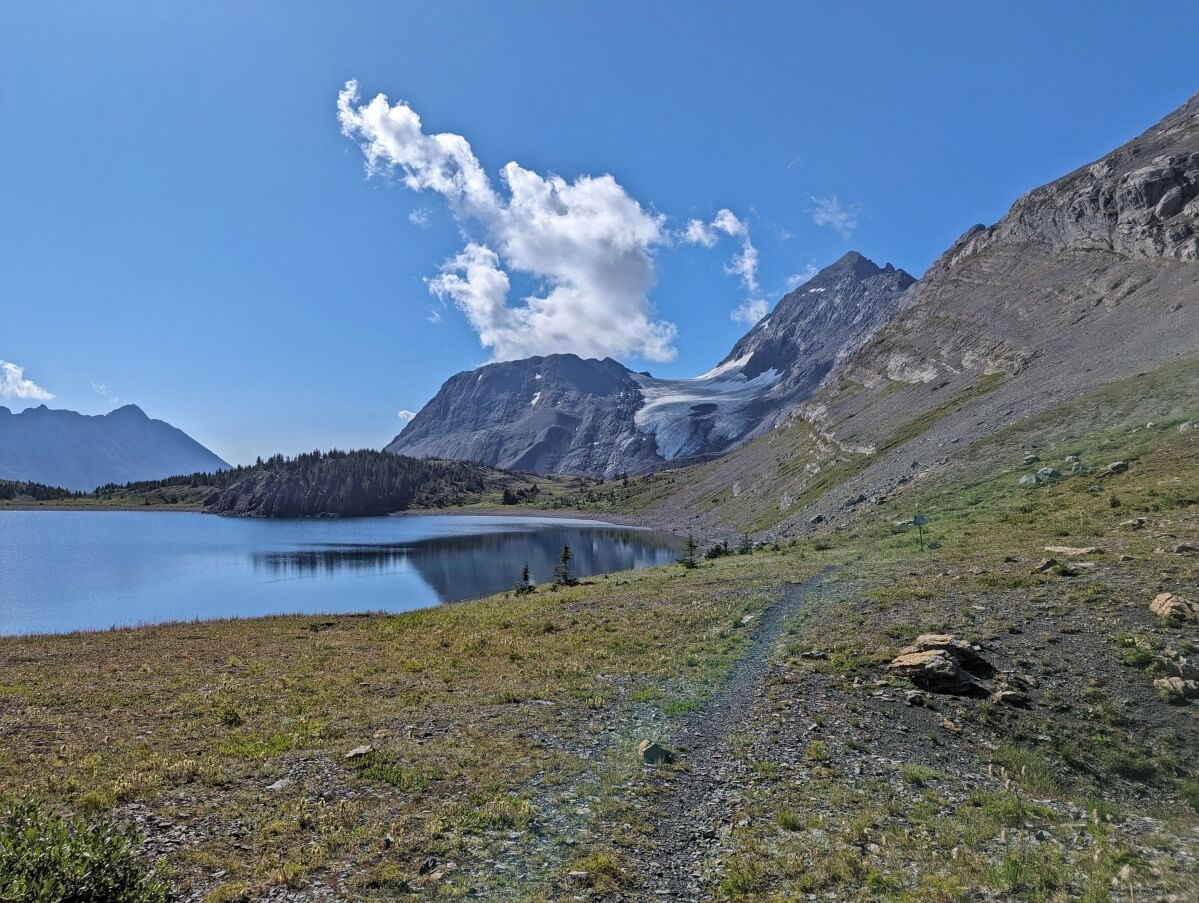 Dirt hiking trail on Great Divide leading past calm lake with glacier behind