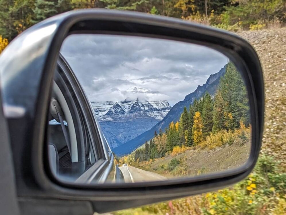 Wing mirror view of Mount Robson in fall, with road stetching out behind, large snowy mountain and mix of yellow/green foliage
