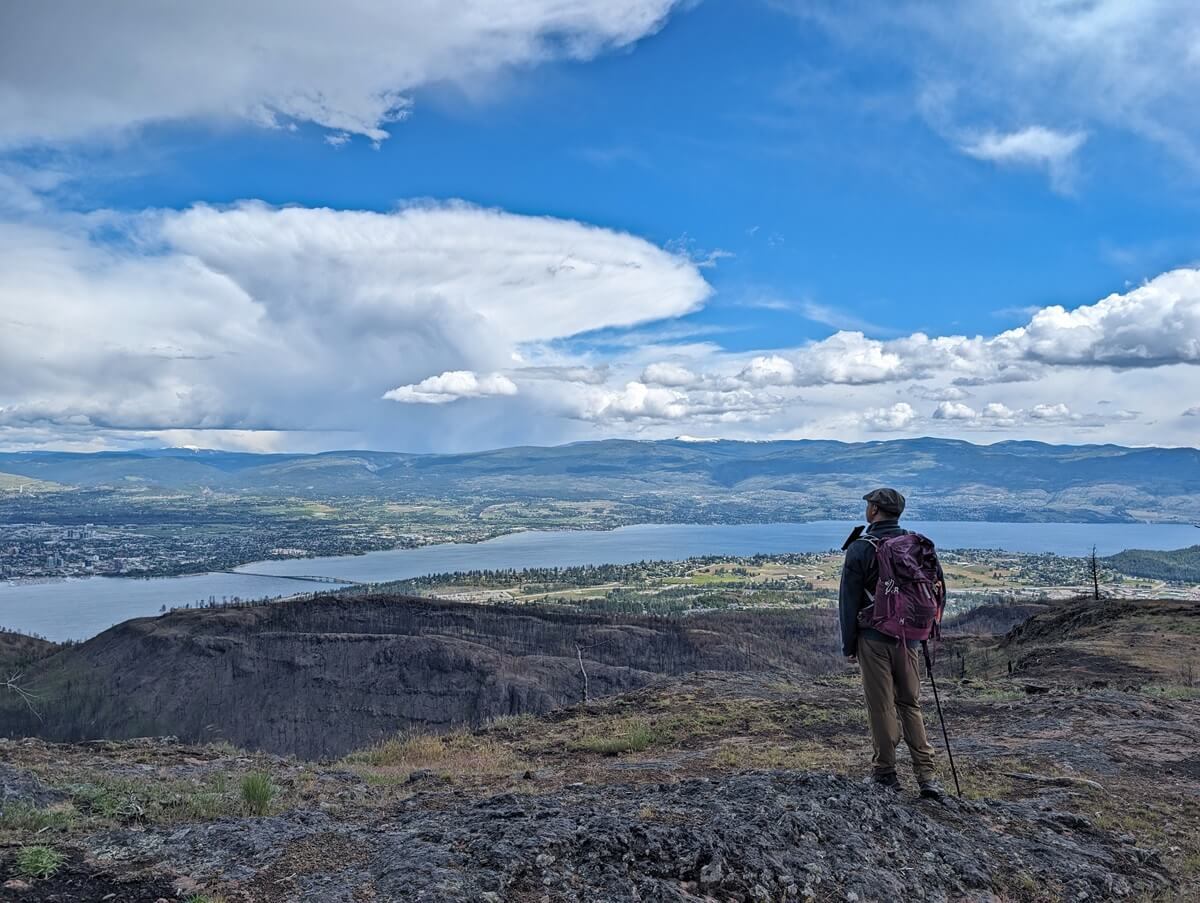 Back view of JR standing on rocky area looking out to view of Okanagan Lake and the city of Kelowna