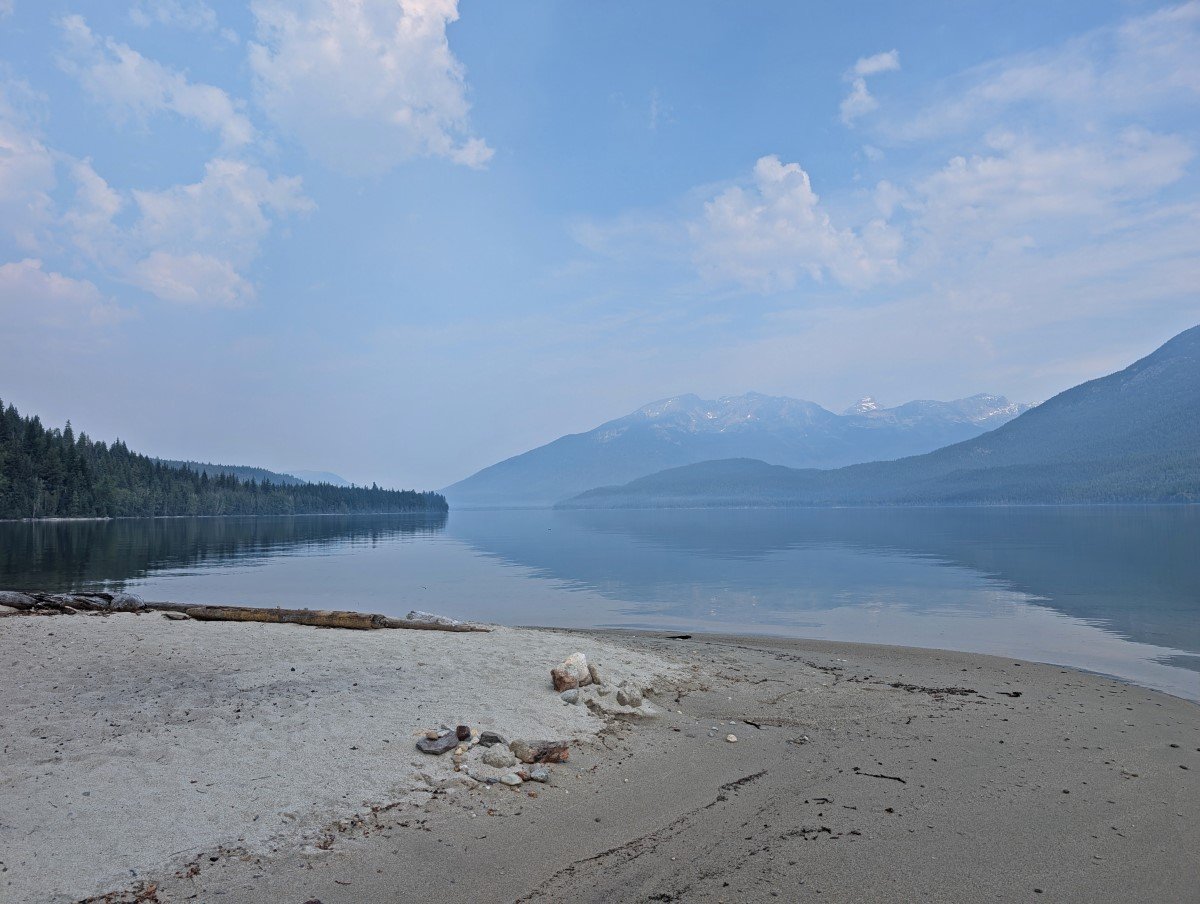 Sandy beach peninsula on calm mirror like lake with mountains visible in the background amidst smoky skies