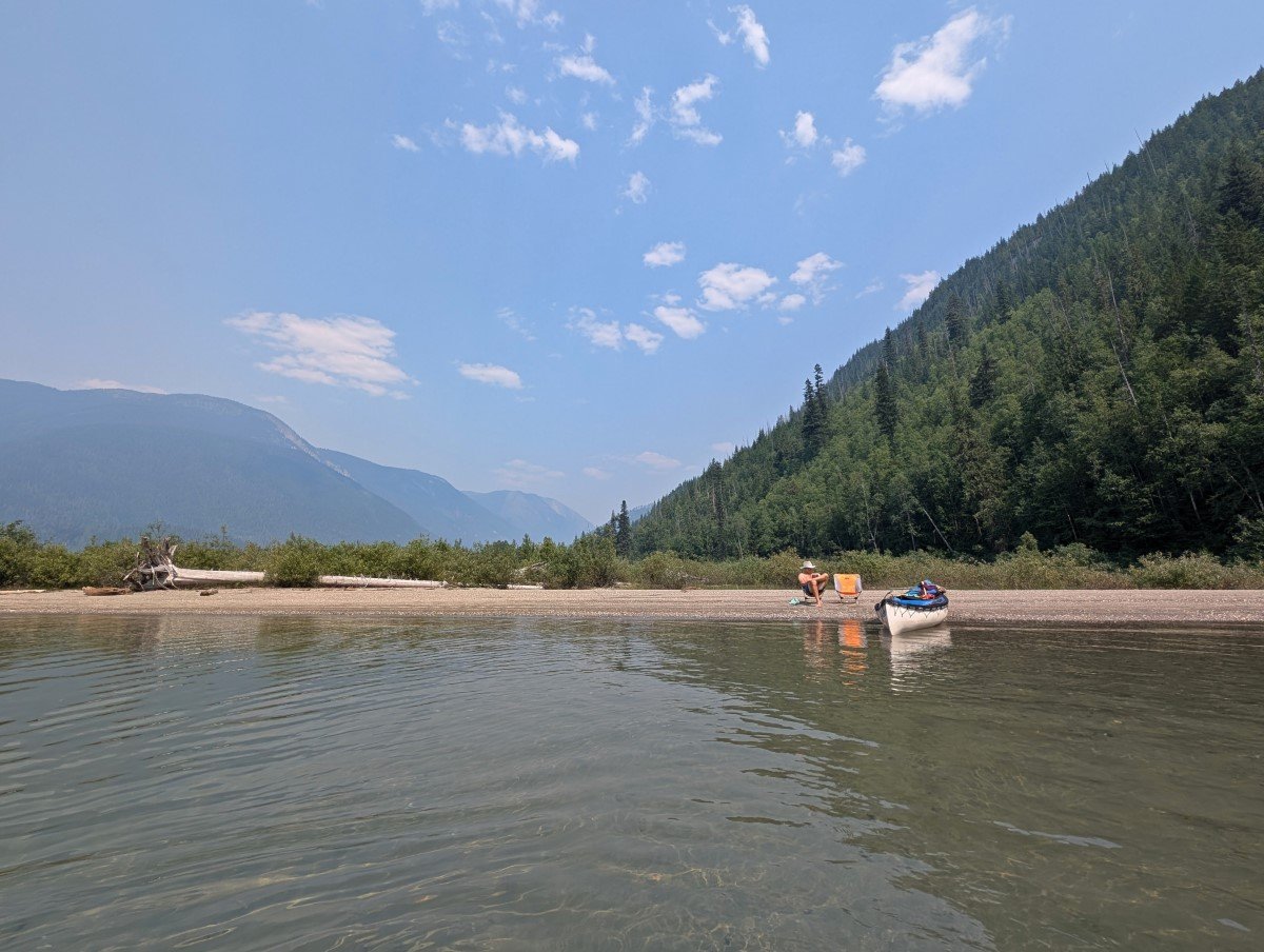 Water view looking back at shore, where a person sits in a chair and a canoe sits on the beach. There are forested mountains behind
