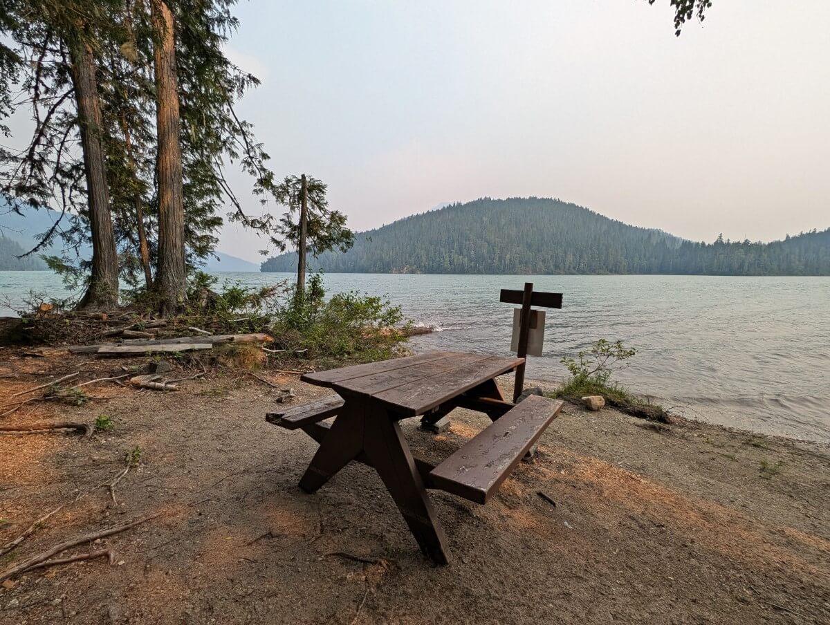A picnic table and sign in an opea area looking over the windy lake. There are some trees on the left and more lining the shoreline of the other side of the lake