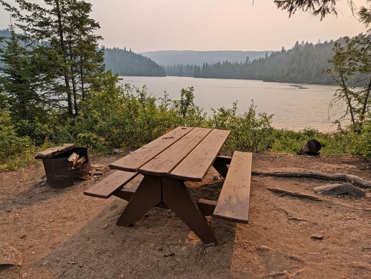 A picnic table and fire pit look out on a high bluff over a calm lake with smoky skies