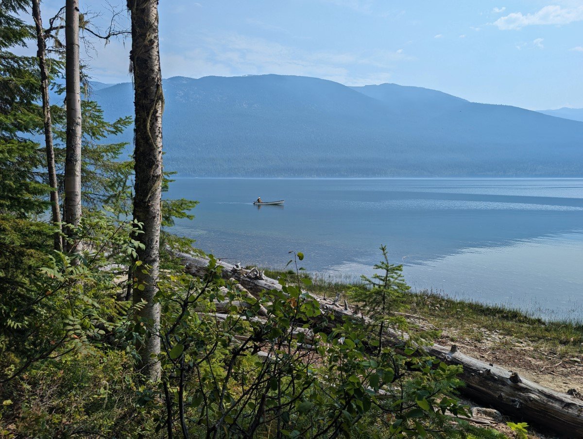 Looking through the trees to calm Clearwater Lake, where a canoeist is paddling in the distance. There are forested mountains on the other side of the lake