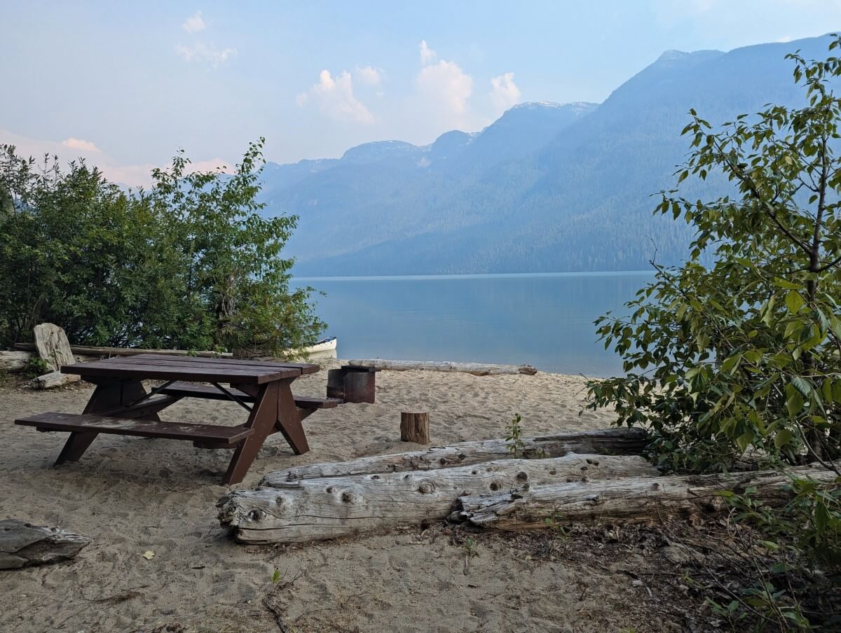 Sandy beach area with picnic table and fire pit in front of a calm lake and mountain view