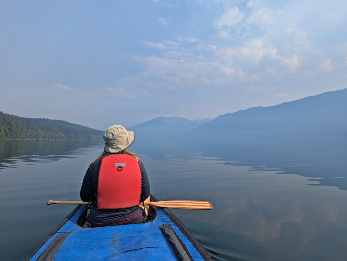 Back view of paddler sat in canoe looking out to the view of the calm lake surrounded by forested mountains