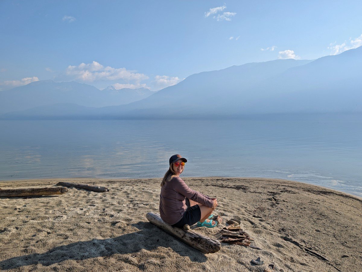 Gemma sits on a log on the beach next to calm Clearwater Lake in Wells Gray