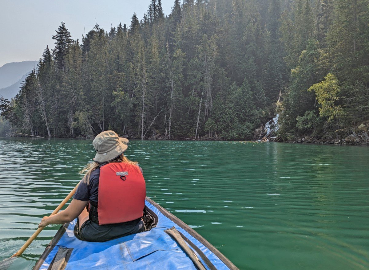 Back view of canoe paddler in canoe on green coloured lake, looking at waterfall on right hand side