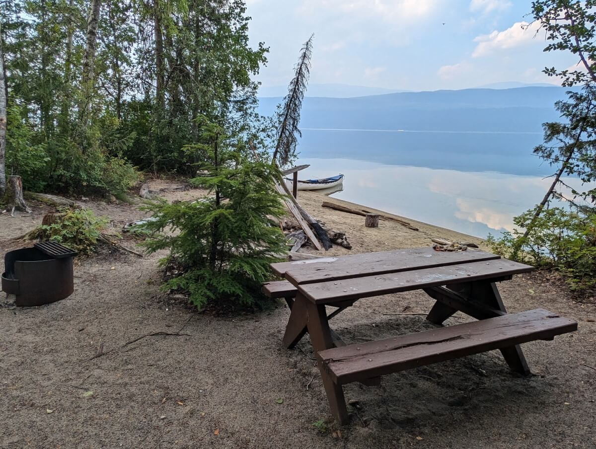 Mostly open campground view with picnic table, fire pit, scattered trees and lake views in background. There is a canoe resting on the shore