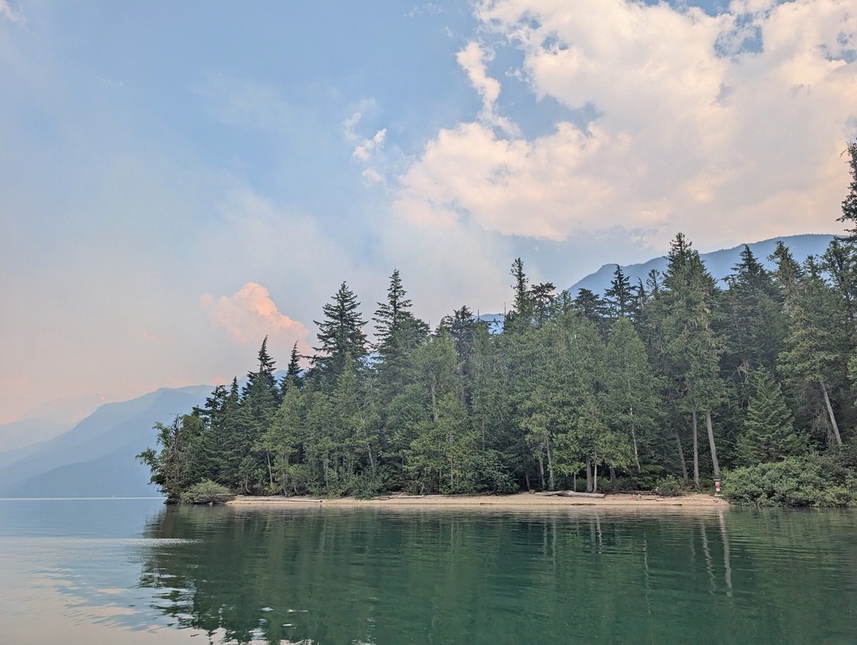Canoe view of a sandy beach from the lake, lined by forest and backdropped by mountains