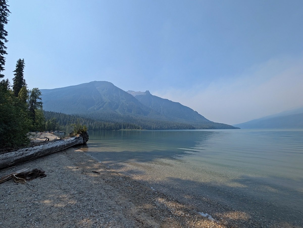 Pebble beach next to calm lake with forested mountains in the background. There is a very large driftwood log on the beach