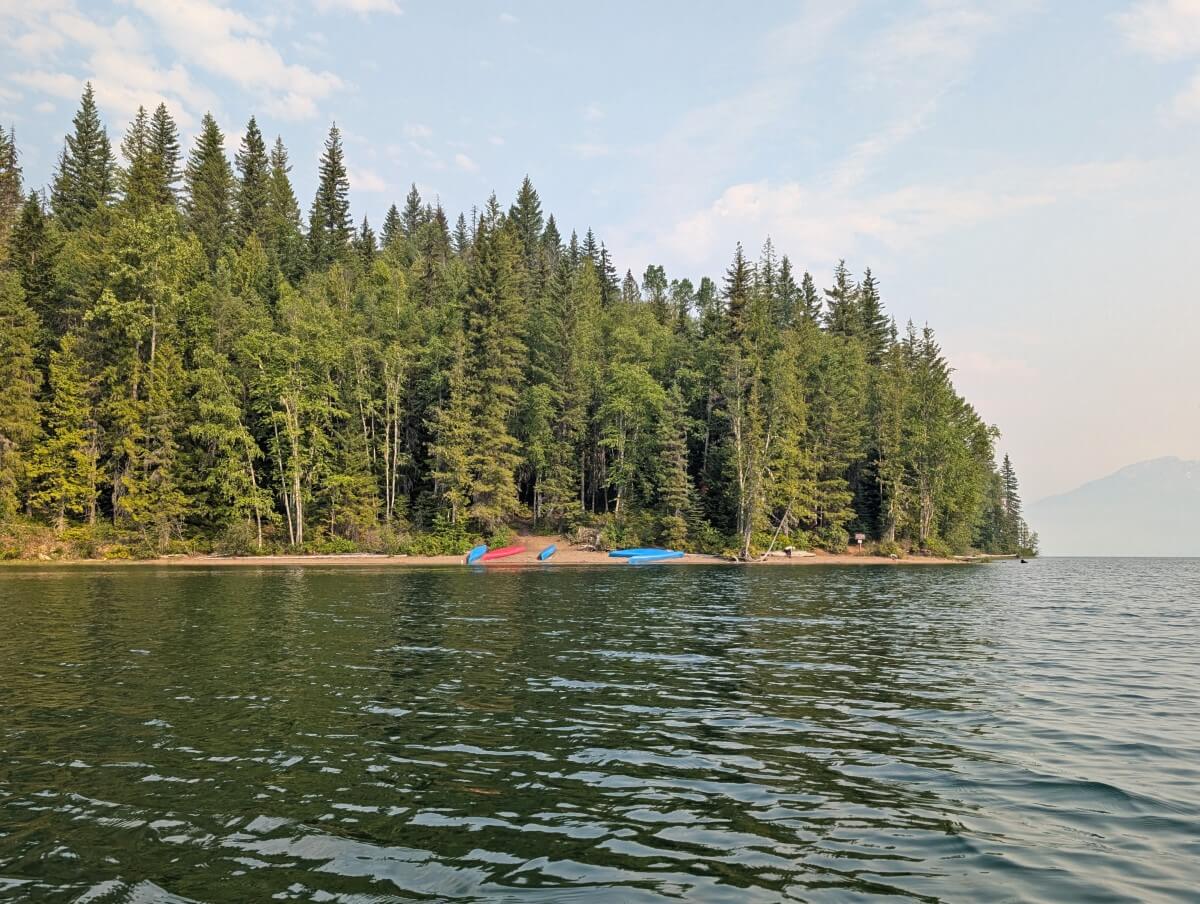 Canoe view of fairly calm lake, looking towards sandy shoreline lined by trees, with five brightly coloured canoes on sandy beach