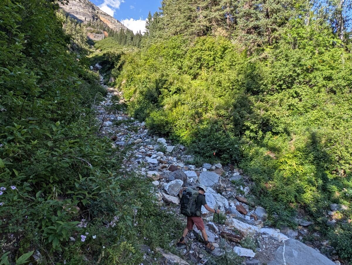 Side view of hiker crossing rocky avalanche chute on Little Peters Lake, with forest on either side