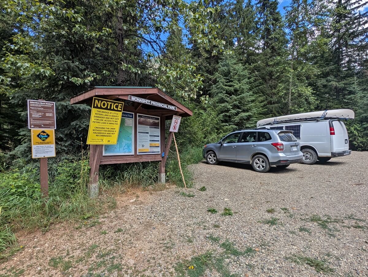 Monashee Provincial Park kiosk and signage with two parked vehicles on the right hand side. There is a large yellow coloured notice posted on the kiosk