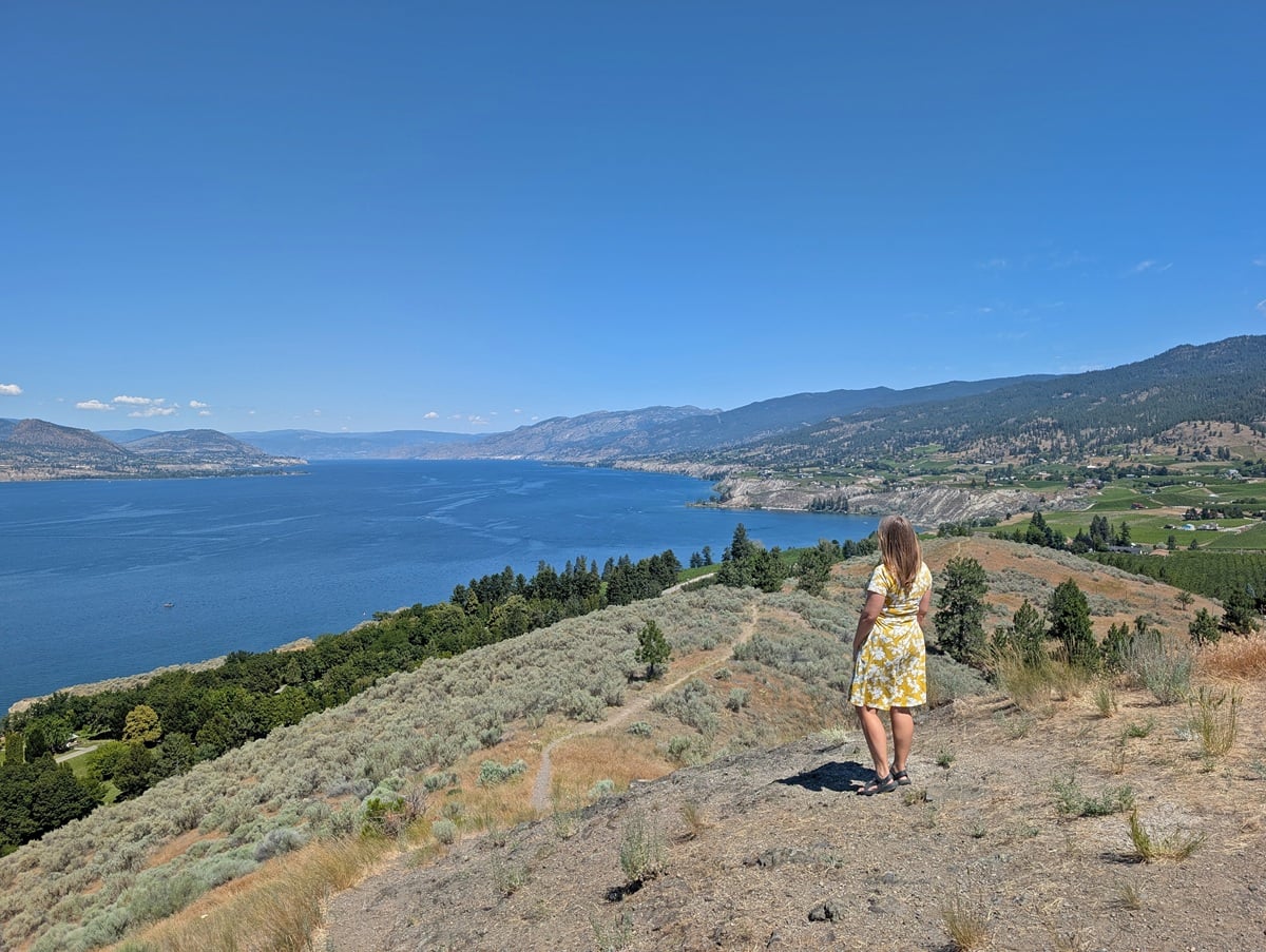 Back view of Gemma in a yellow dress looking out to the lake and vineyard views from Munson Mountain in Penticton