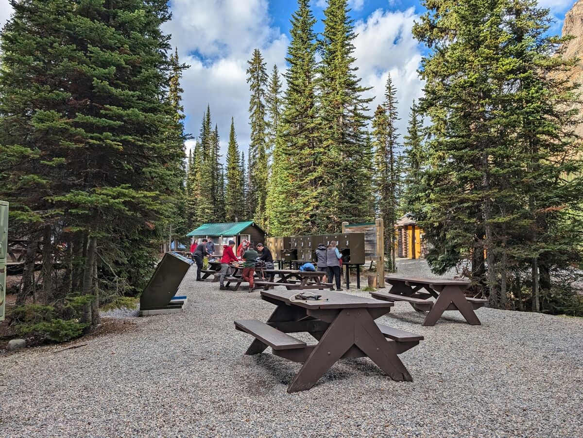 Wooden picnic tables on gravel surface in the Lake O'Hara campground. There are trash bins to the left and food lockers on the right. Trees surround the communal space. There are hikers preparing backpacks