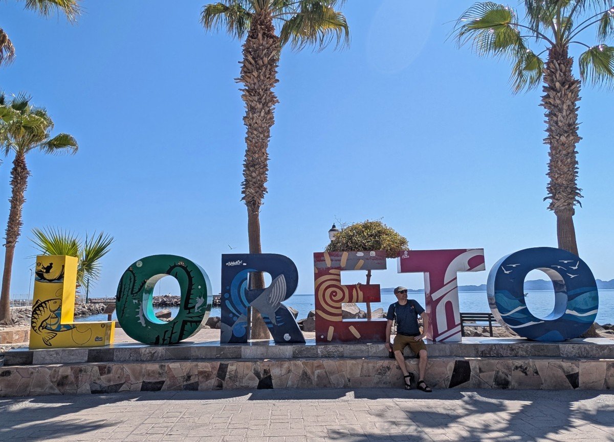 JR sat on colourful Loreto sign in front of palm trees and ocean in Mexico