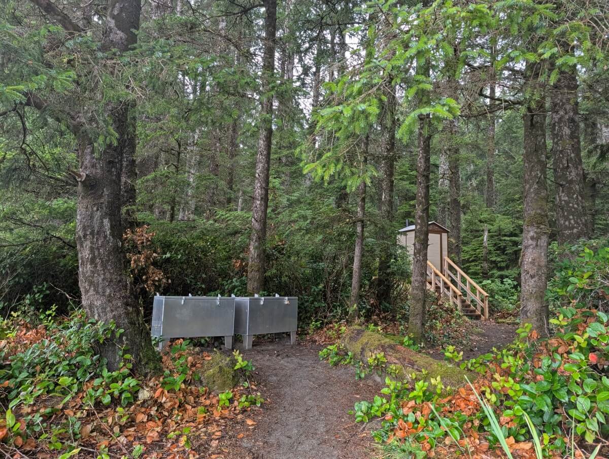 Two metal food caches in the forest next to an outhouse, accessible by steps