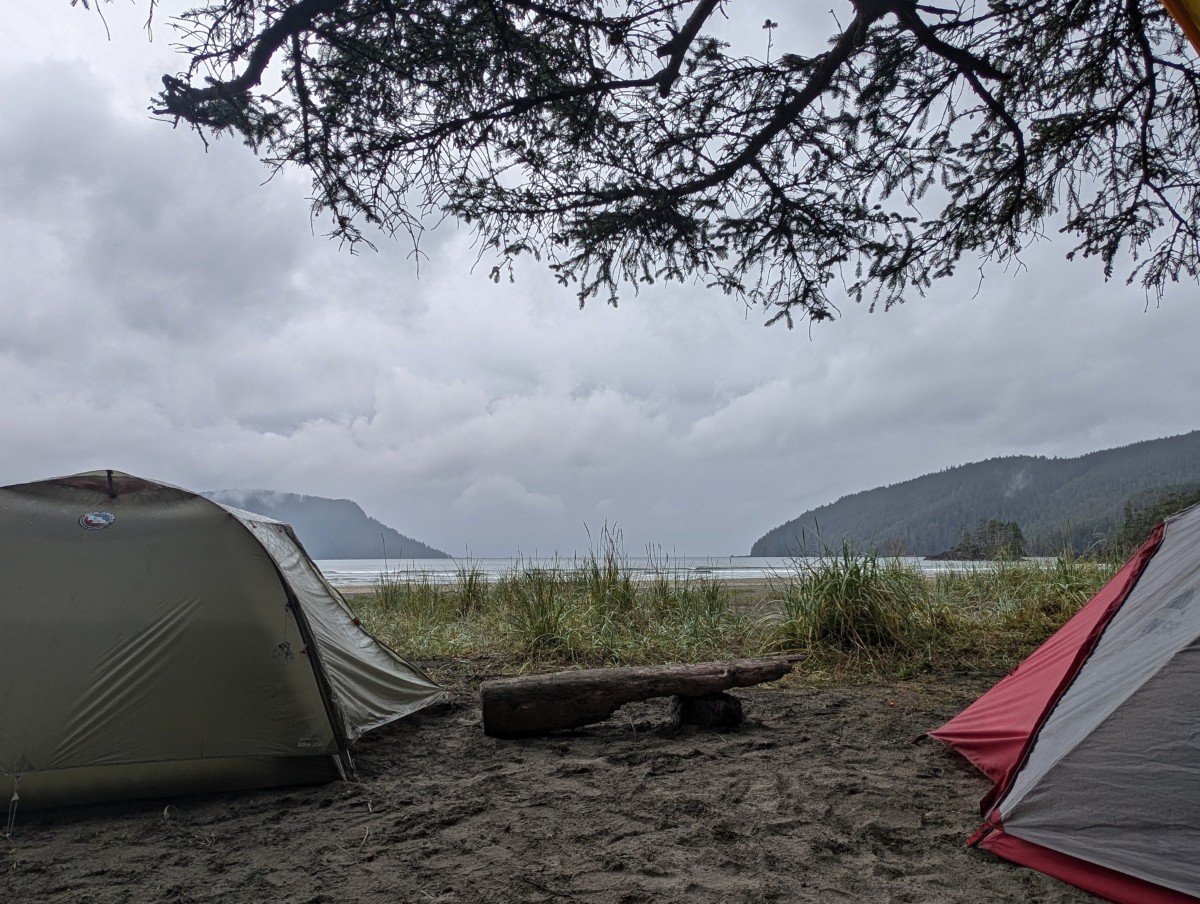 Looking through two tents set up on San Josef Bay beach, with a log bench inbetween. The ocean is visible beyond the grass in front of the tents