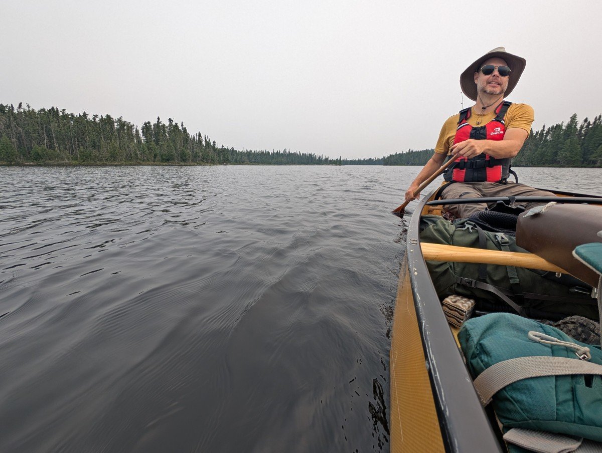 Side view of JR paddling a yellow canoe in Wabakimi Provincial Park, with calm lake to left, lined by forest