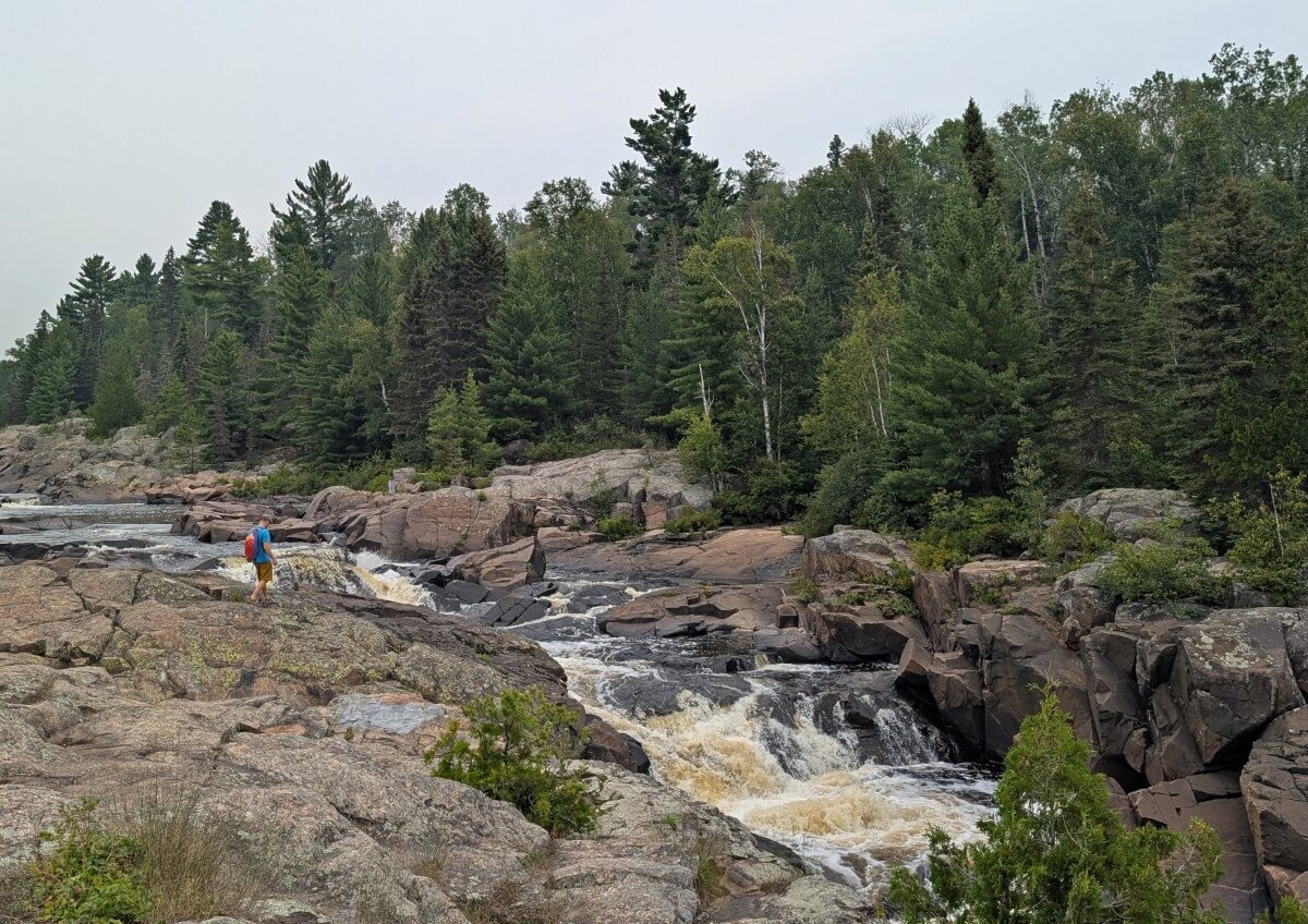 Series of rapids in Cascades Conservation Area surrounded by rusty coloured rocks. A hiker is apporoaching the edge of the river, which is lined by forest on the other side
