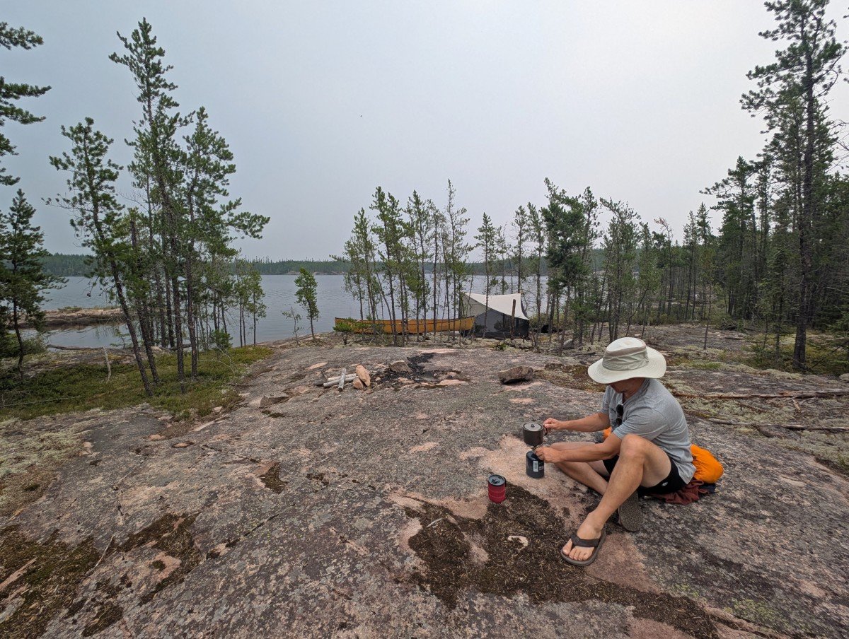 JR sits on the ground adjusting a camp stove, with bug tent and canoe in background at campsite, next to lake