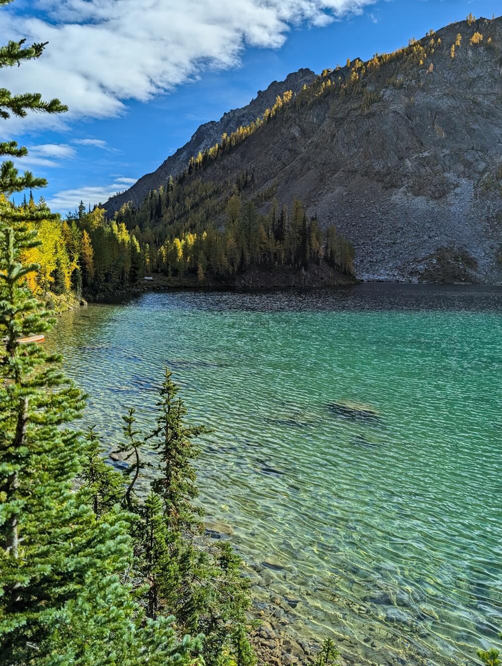 Side view of turquoise Diana Lake from lakeshore trail with trees bordering the edge and rugged rock walls rising above