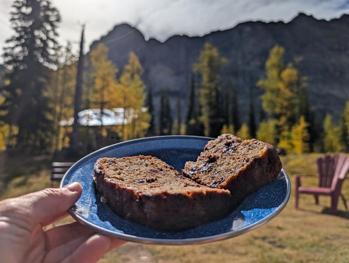 A hand holds up a plate of cake at Diana Lake Lodge, with golden larch trees in the background