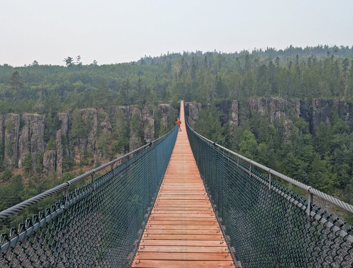 Suspension bridge view towards lone hiker in orange shirt leaning on the side of the bridge, with tall rock cliffs on the other side of the canyon