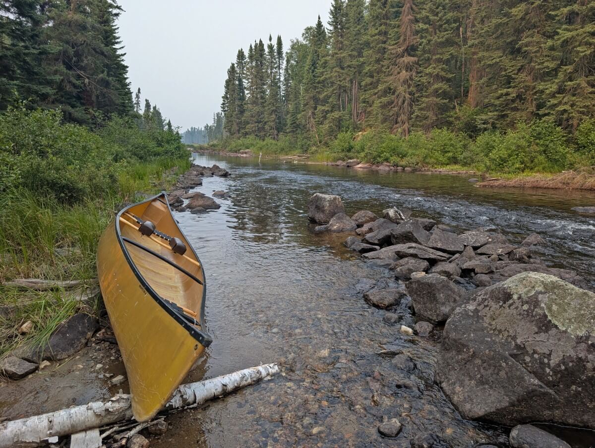 A canoe rests on the left shore of a river in Wabakimi Provincial Park. The river is lined by trees