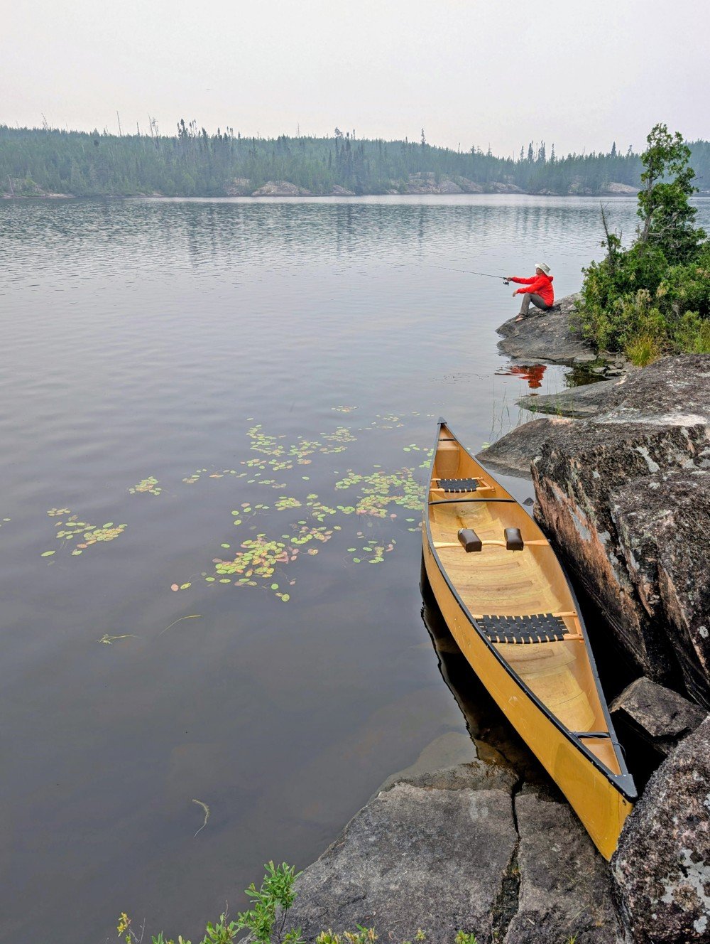 A yellow canoe rests on a calm lake next to a rock bluff, at the back of which someone is sitting fishing