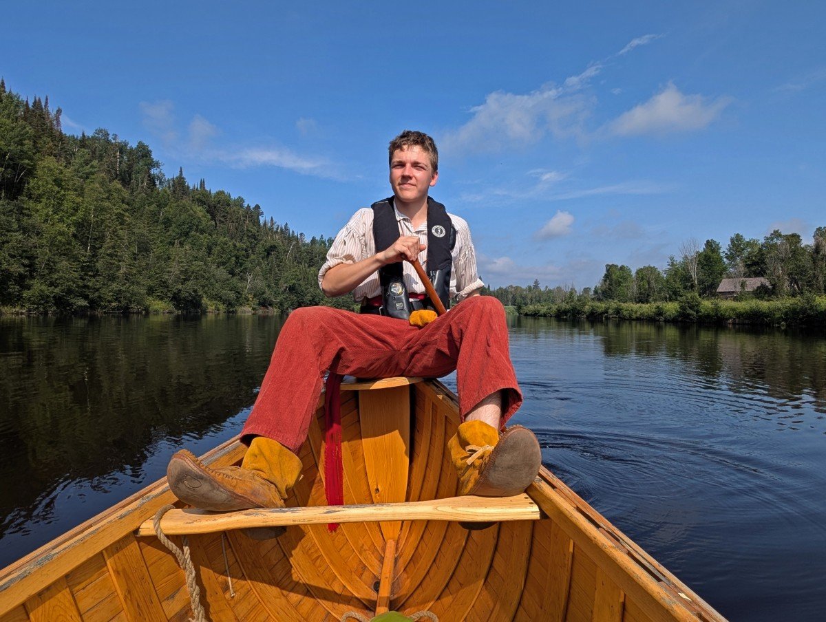 Looking behind on a large voyageur canoe on the river to a man holding a paddle and sitting at the very back of the canoe, wearing 19th century clothing
