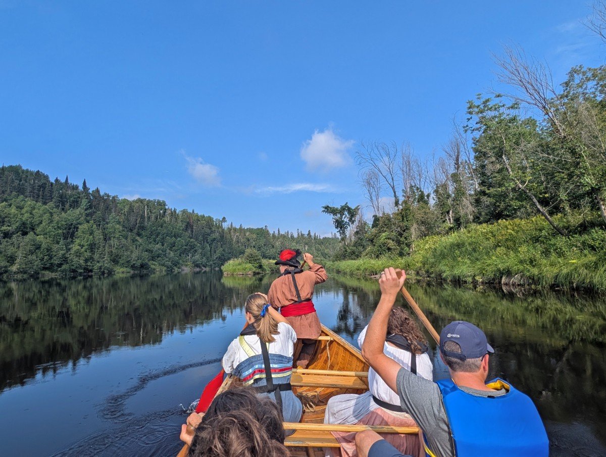 Back view of five paddlers in voyageur canoe on camp river near Fort William Historical Park. Three of the paddlers are in 19th century costume