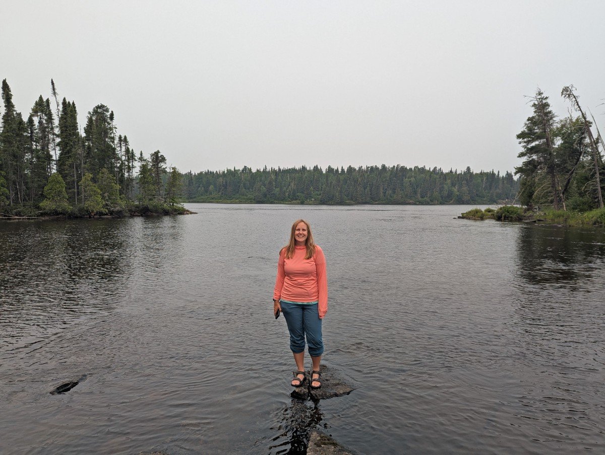 Gemma stands on a rock seemingly in the middle of a lake in Wabakimi Provincial Park, with trees visible behind