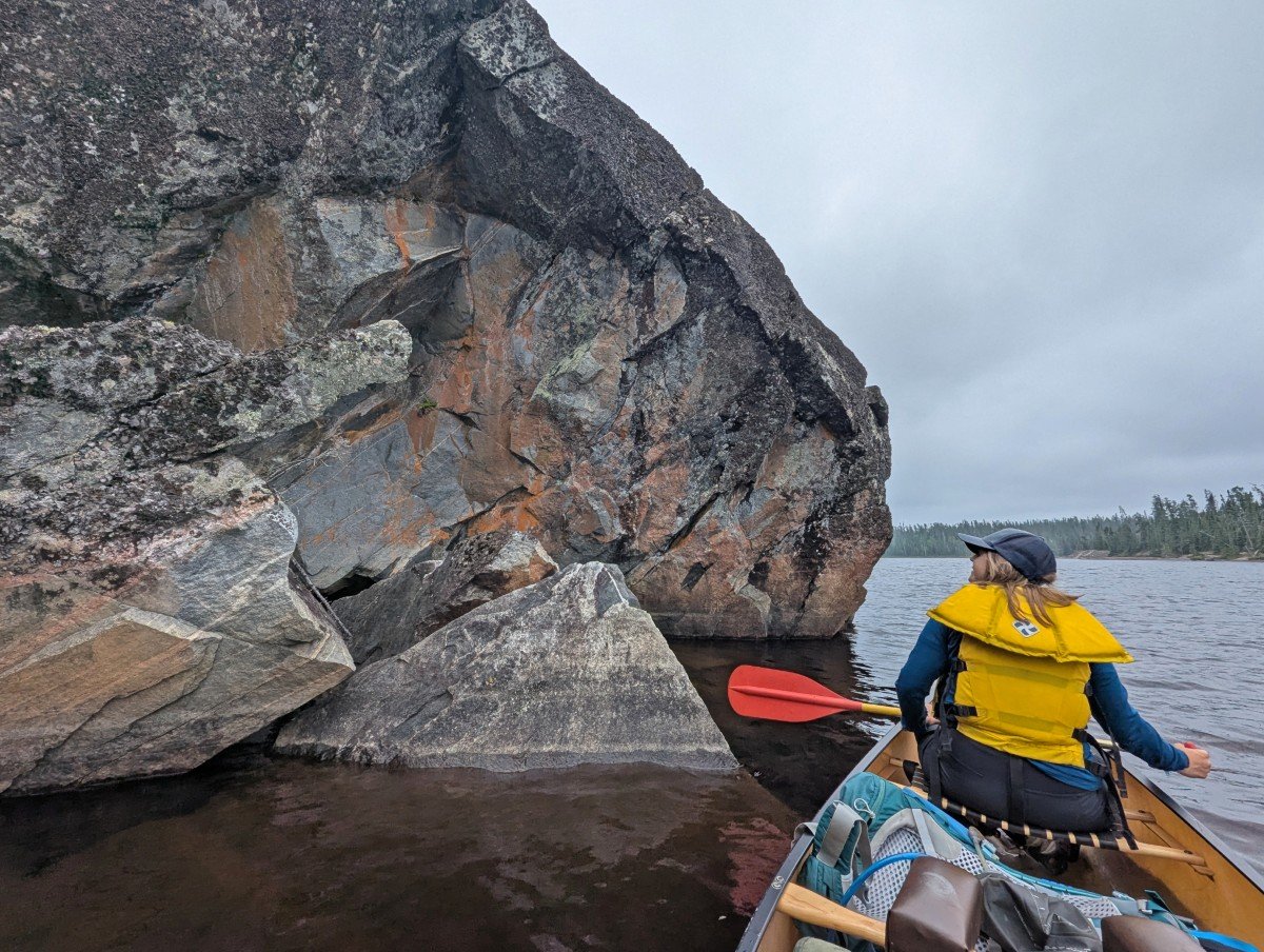 Back view of Gemma sitting in canoe looking up at colourful rock cliff on left