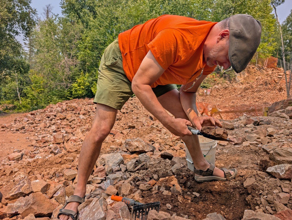 JR bends over in amethyst quarry with rock on shovel, looking for gems
