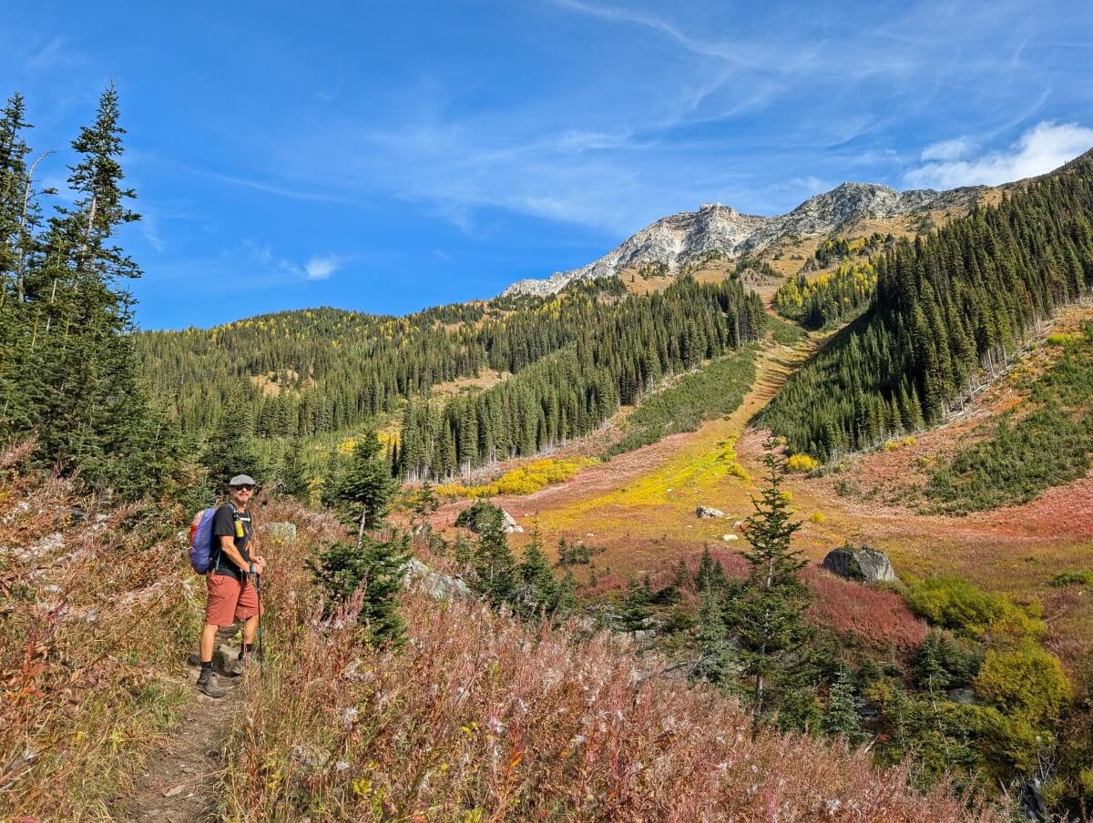 Side view of JR on left hand side of trail through high foliage area on the way to Diana Lake. There are colourful mountain slope views on the right hand side, with patches of forest