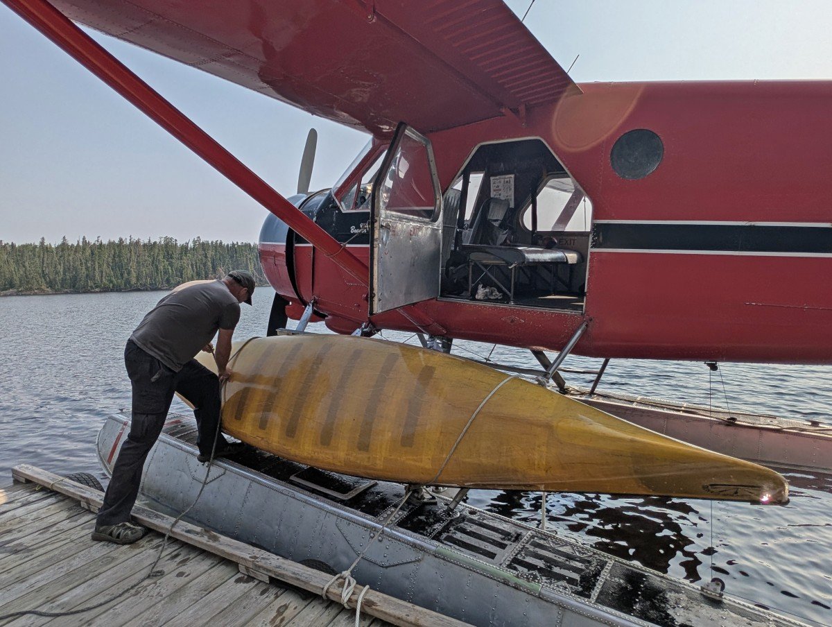 A man loads a yellow canoe onto the side of a red floatplane, parked on a lake