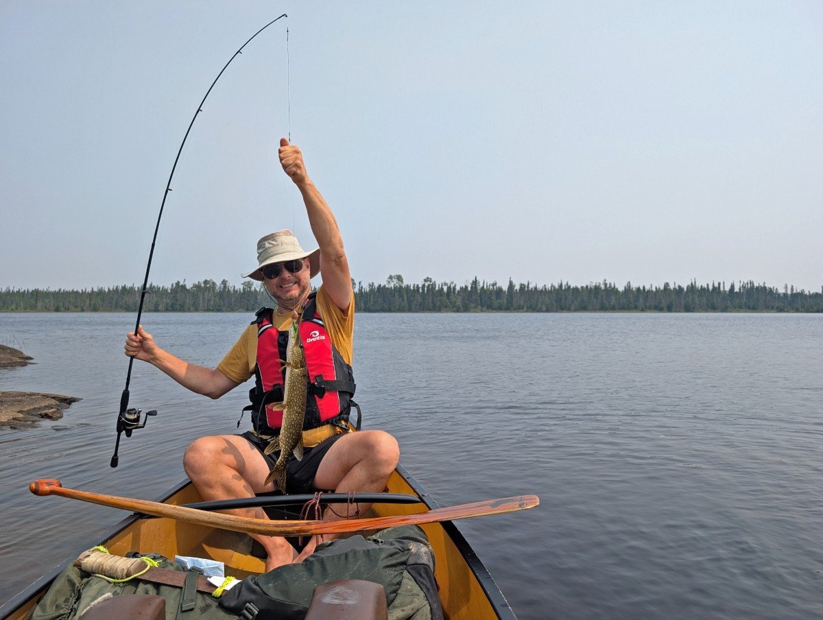 JR sits in canoe on a calm Wabakimi lake, holding up fishing line with northern pike on end of line