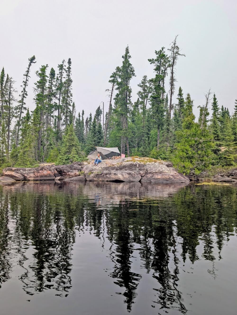 Water view of campsite in Wabakimi Provincial Park, featuring set up tent and tarp structure above rock bluff, in front of forest. There is a person sat on the rock