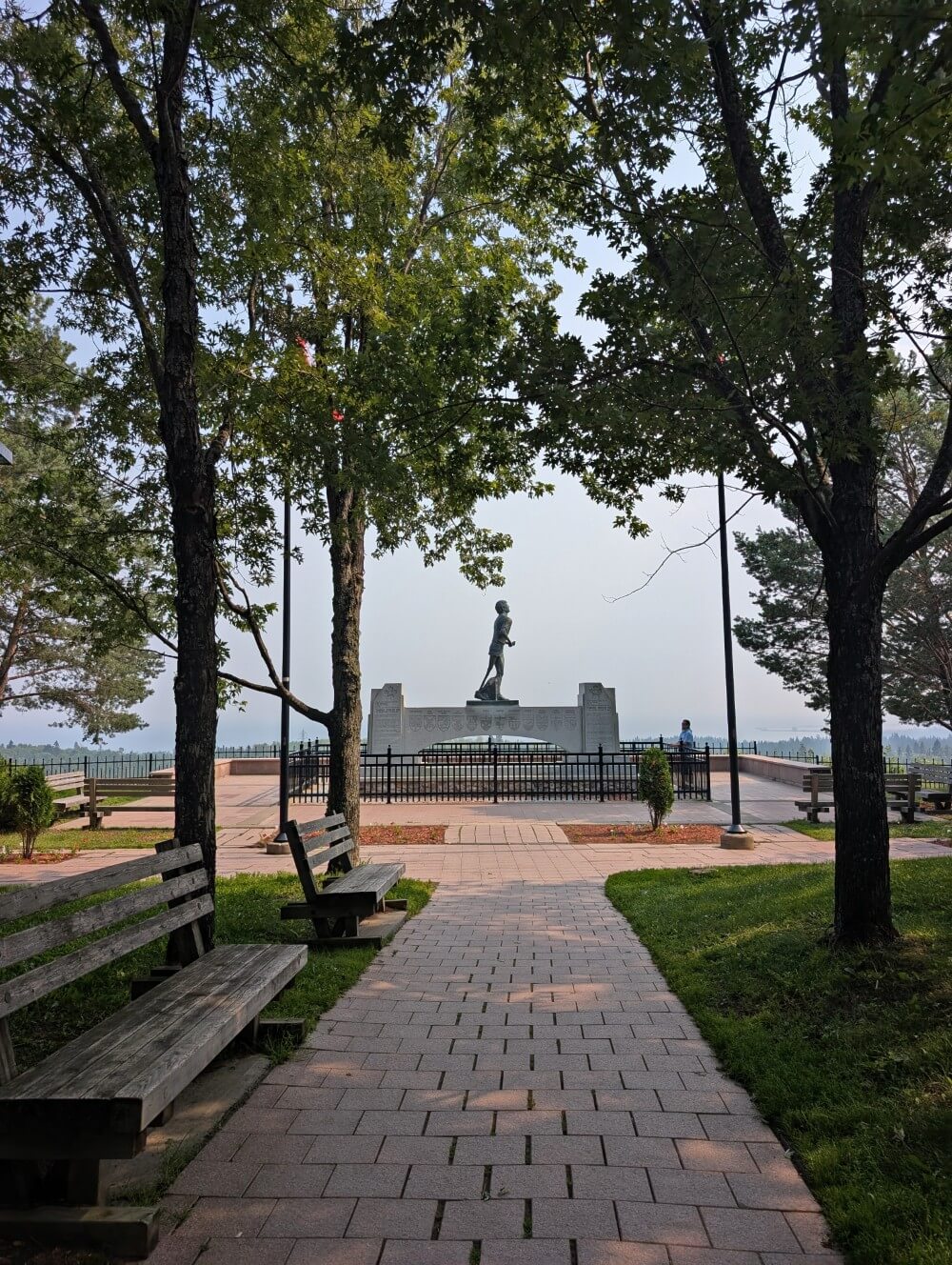 Looking ahead through the trees to the fenced Terry Fox monument, featuring a larger than life human figure