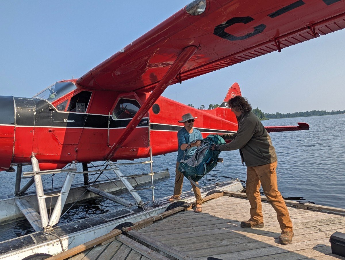 Two men pass a large backpack between them on a dock next to a red floatplane