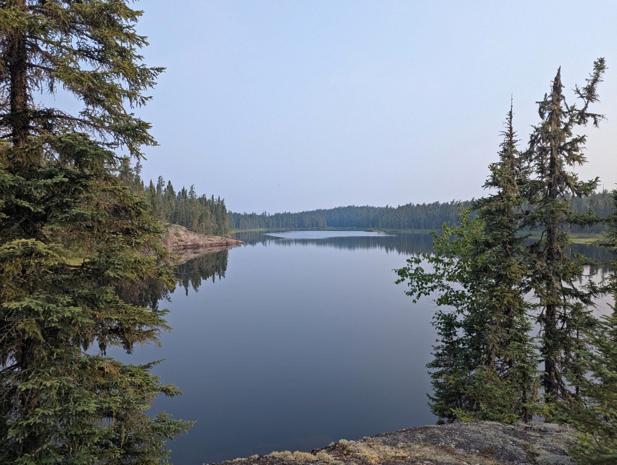 Campsite view in Wabakimi, looking out between trees to perfectly still lake below