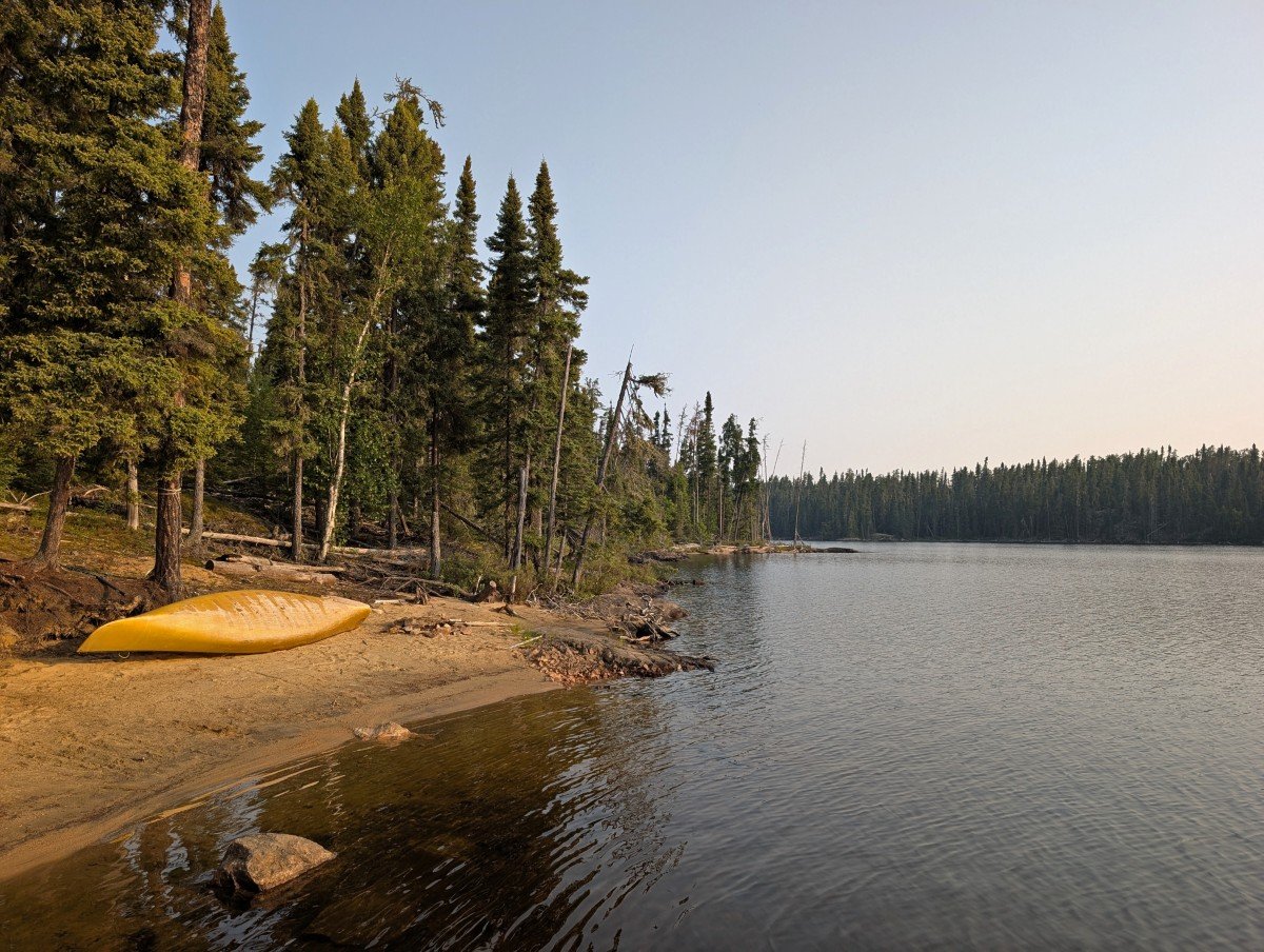 A yellow canoe is upside down on a sandy beach next to a lake, in front of forest