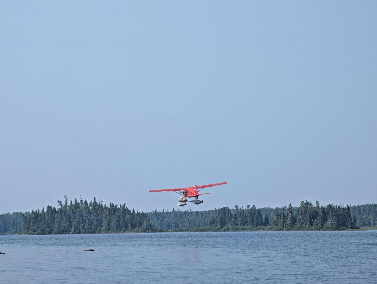 A red floatplane is flying away above the lake with a canoe strapped on the side