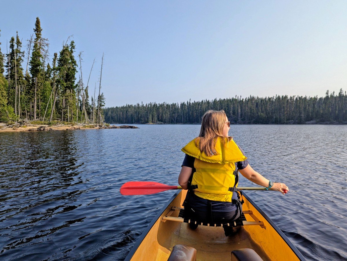 Back view of paddler in canoe in Wabakimi Provincial Park, with forested shoreline ahead and to the left. The lake is calm